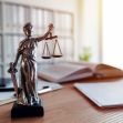 A statue of Lady Justice holding scales and a sword on a desk with legal books and documents in the background. A statue of Lady Justice holding scales and a sword on a desk with legal books and documents in the background.
