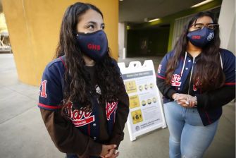 Two students wearing masks and school jackets stand outside a building with a sign in the background.