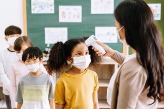 A teacher checking the temperature of a student in a classroom while other students wait in line, all wearing masks.