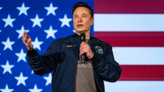 Elon Musk speaking at an event with a U.S. flag backdrop.