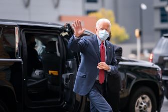 Joe Biden waving while exiting a vehicle, wearing a suit and mask.
