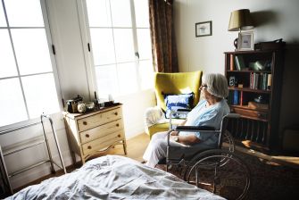An elderly woman in a wheelchair looking out a window in a nursing home room.