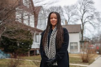 A woman standing outdoors in a neighborhood, wearing a black coat and patterned scarf.