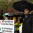 A man holding an umbrella stands in front of signs expressing opposition to the Cultural Competence Action Plan (CCAP) and critical race theory during a demonstration. A man holding an umbrella stands in front of signs expressing opposition to the Cultural Competence Action Plan (CCAP) and critical race theory during a demonstration.