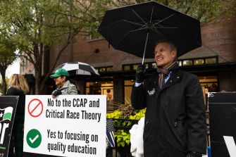 A man holding an umbrella stands in front of signs expressing opposition to the Cultural Competence Action Plan (CCAP) and critical race theory during a demonstration.