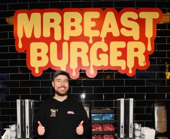 A person standing in front of a "MrBeast Burger" sign, smiling and giving a thumbs up.