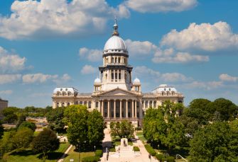 The Illinois State Capitol building in Springfield, surrounded by trees and a blue sky with clouds.