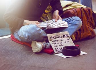 A person sitting on the ground with a sign that reads about being homeless and asking for help, surrounded by papers and belongings.
