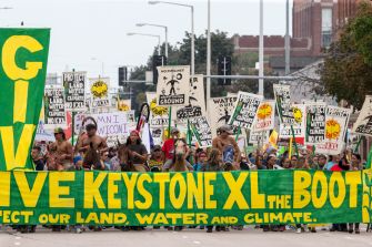 A protest march with participants holding banners and signs advocating against the Keystone XL Pipeline, emphasizing the protection of land, water, and climate.
