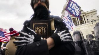 A person holding a Bible while participating in a rally at the Capitol, surrounded by flags and police officers in riot gear.