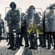 A line of police in riot gear holding shields during a protest. A line of police in riot gear holding shields during a protest.