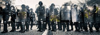 A line of police in riot gear holding shields during a protest.
