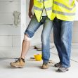 An injured construction worker with a bandaged knee, assisted by another individual on a worksite. An injured construction worker with a bandaged knee, assisted by another individual on a worksite.