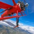 A person hangs from the wing of a red bi-plane, soaring high above scenic landscapes under a clear blue sky. A person hangs from the wing of a red bi-plane, soaring high above scenic landscapes under a clear blue sky.