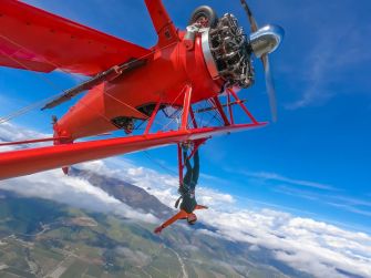 A person hangs from the wing of a red bi-plane, soaring high above scenic landscapes under a clear blue sky.
