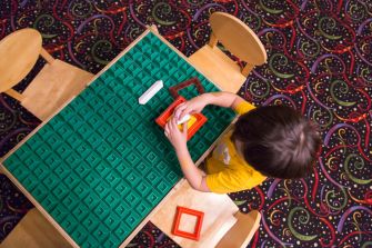 A top-down view of a young child playing with colorful building blocks on a textured green table, surrounded by empty wooden chairs.