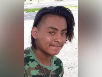 A young man with long hair and a camouflage shirt smiles at the camera, standing outdoors.