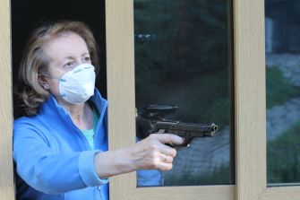 A woman wearing a mask points a handgun out of a window.