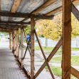 Sidewalk shed structure covering a walkway with wooden supports and a roof, surrounded by autumn foliage and parked cars in the background. Sidewalk shed structure covering a walkway with wooden supports and a roof, surrounded by autumn foliage and parked cars in the background.