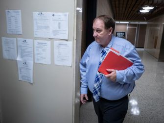 A man in a blue dress shirt and plaid tie walks past a wall covered with official documents and notices.