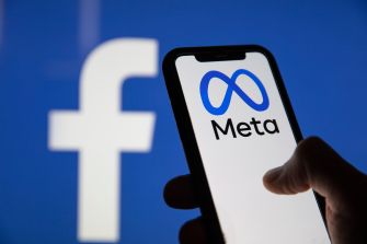 A person holding a smartphone displaying the Meta logo, with the Facebook logo visible in the background.