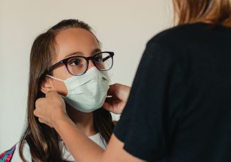 A child wearing a mask while being helped by an adult, highlighting safety measures in response to COVID-19.