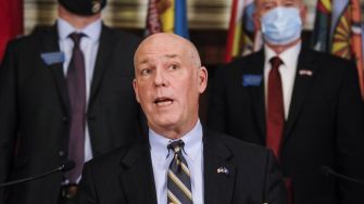 A man in a suit speaks at a press conference, with two individuals in the background, one wearing a mask. The setting includes flags hanging on the wall.