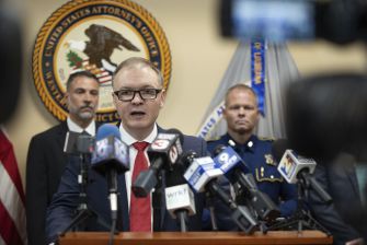 A press conference regarding the indictment of law enforcement officials and a business owner for U-Visa fraud, with a U.S. Attorney speaking surrounded by law enforcement representatives.