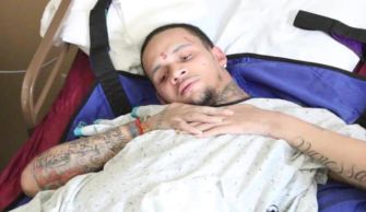 A man lying in a hospital bed, appearing to be in a medical facility, with visible tattoos and a concerned expression.