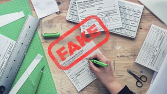 A hand writing on a COVID-19 vaccination card with various tools and papers on a table, marked with the word "FAKE" in red.