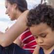 A woman sits beside a young boy, who appears to be lost in thought, while they are outdoors. The woman wears a red and white striped top, and the boy has curly hair. A woman sits beside a young boy, who appears to be lost in thought, while they are outdoors. The woman wears a red and white striped top, and the boy has curly hair.