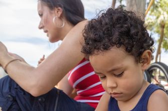 A woman sits beside a young boy, who appears to be lost in thought, while they are outdoors. The woman wears a red and white striped top, and the boy has curly hair.