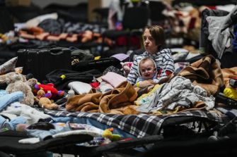 A woman sitting on a makeshift bed surrounded by blankets and luggage, holding a baby, in a crowded shelter setting.