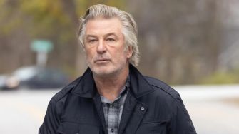 Alec Baldwin walking outside with a serious expression, wearing a black jacket over a plaid shirt.