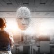 A person standing in an office environment, looking at a digital representation of a human face made up of particles and light. A person standing in an office environment, looking at a digital representation of a human face made up of particles and light.