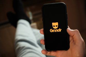 Image of a smartphone displaying the Grindr logo while a person in casual clothing holds it, with their legs visible in the background.