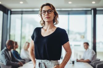 A confident female lawyer standing in a modern office setting, with colleagues engaged in discussion in the background.