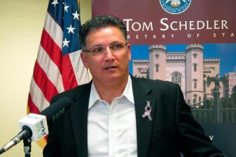A man in a suit speaking at a press conference, with a flag and a backdrop featuring the Louisiana state emblem.