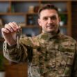 A service member in military camouflage holds a set of keys in a home setting. A service member in military camouflage holds a set of keys in a home setting.