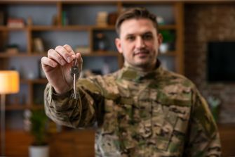 A service member in military camouflage holds a set of keys in a home setting.