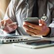 A medical professional using a smartphone and laptop while holding a pen, in a clinical setting. A medical professional using a smartphone and laptop while holding a pen, in a clinical setting.