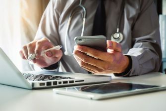 A medical professional using a smartphone and laptop while holding a pen, in a clinical setting.