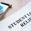 Document labeled "STUDENT LOAN RELIEF" with a pen and notebooks on a wooden surface. Document labeled "STUDENT LOAN RELIEF" with a pen and notebooks on a wooden surface.