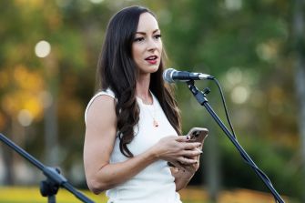 A woman speaking at a microphone outdoors, holding a phone in her hand.