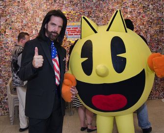 A man in a suit poses with a large, cartoonish Pac-Man character at an event, giving a thumbs up.