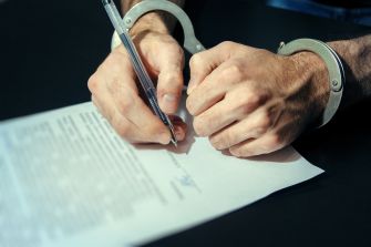 Hands in handcuffs holding a pen above a legal document.