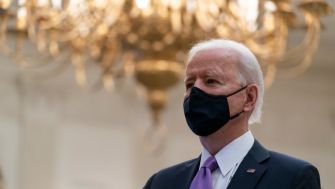 Joe Biden speaking during an event, wearing a mask, with a chandelier in the background.