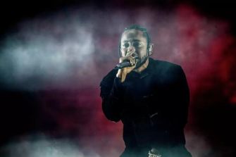 Kendrick Lamar performing on stage with a microphone in hand, surrounded by smoke and dramatic lighting.
