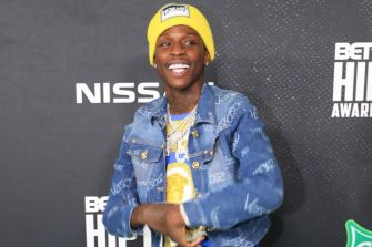 Rapper Quando Rondo smiling at a red carpet event, wearing a yellow beanie and denim jacket.