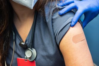 A close-up of a person receiving a vaccination, showing their arm with a bandage and a healthcare professional's gloved hand administering the shot.